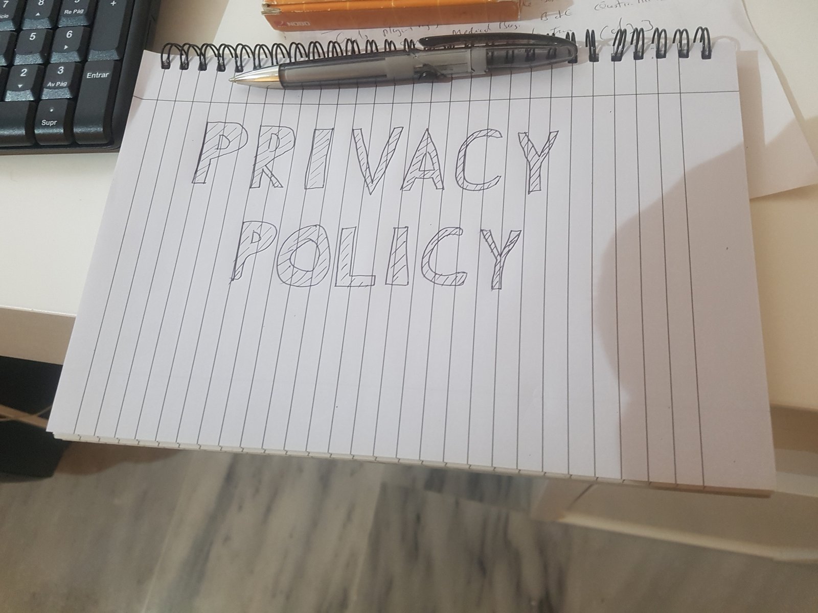 Image shows the word "Privacy Policy" handwritten on a spiral pad with the pen that did it positioned next to a keyboard.